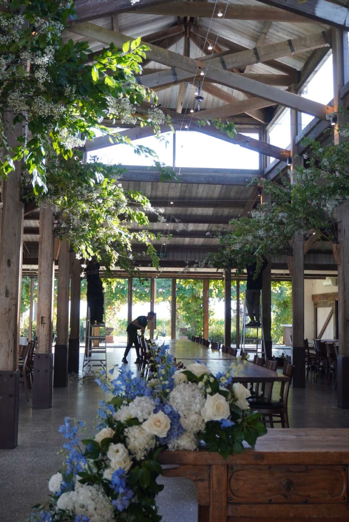 A spacious event venue with wooden beams, large windows, and greenery, featuring a floral arrangement in the foreground and staff preparing the space.