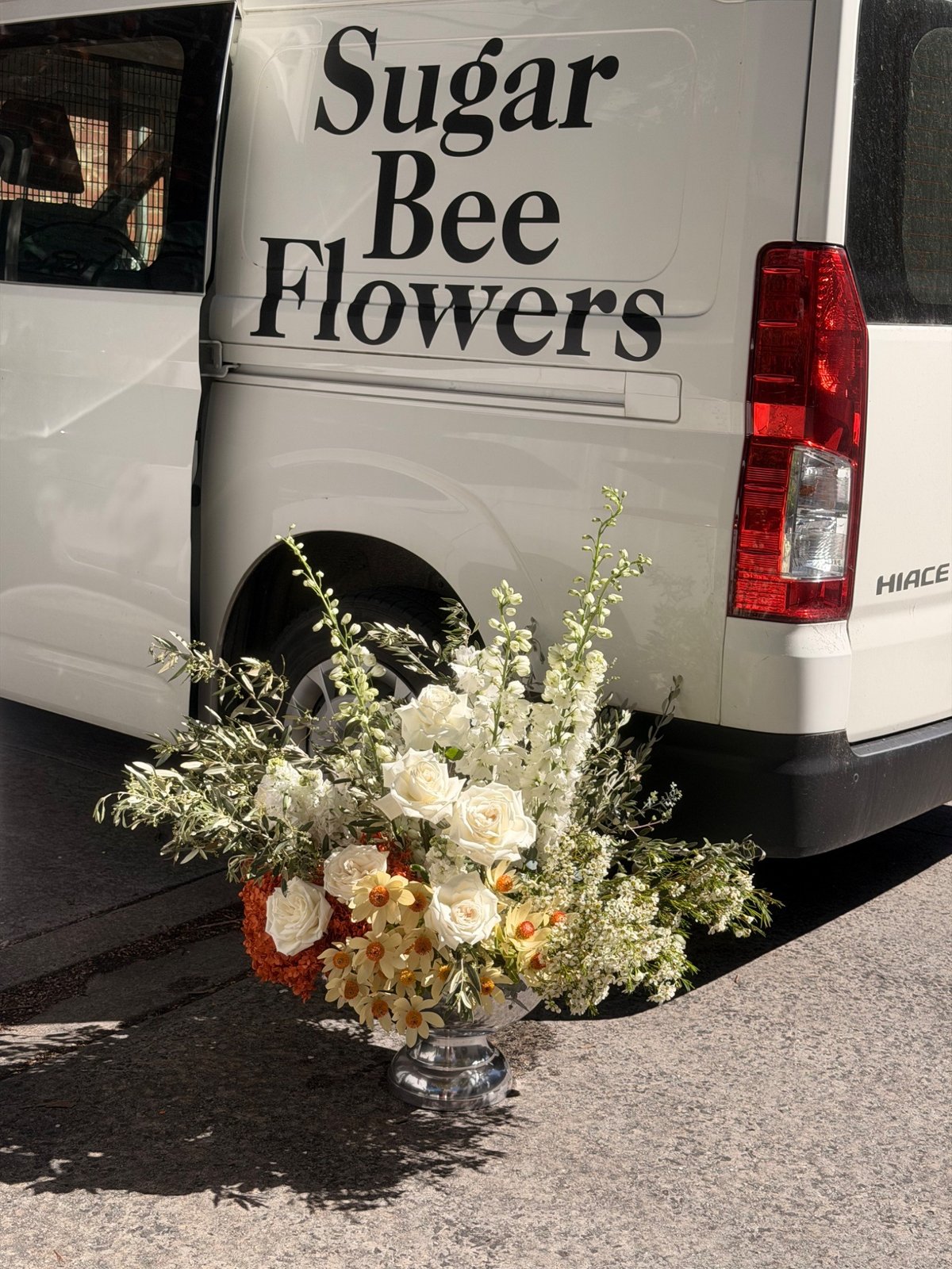 Orange and white event flowers with accents of olive in a silver urn, beside the Sugar Bee Flowers van before the walk across the bridge to Afloat Bar