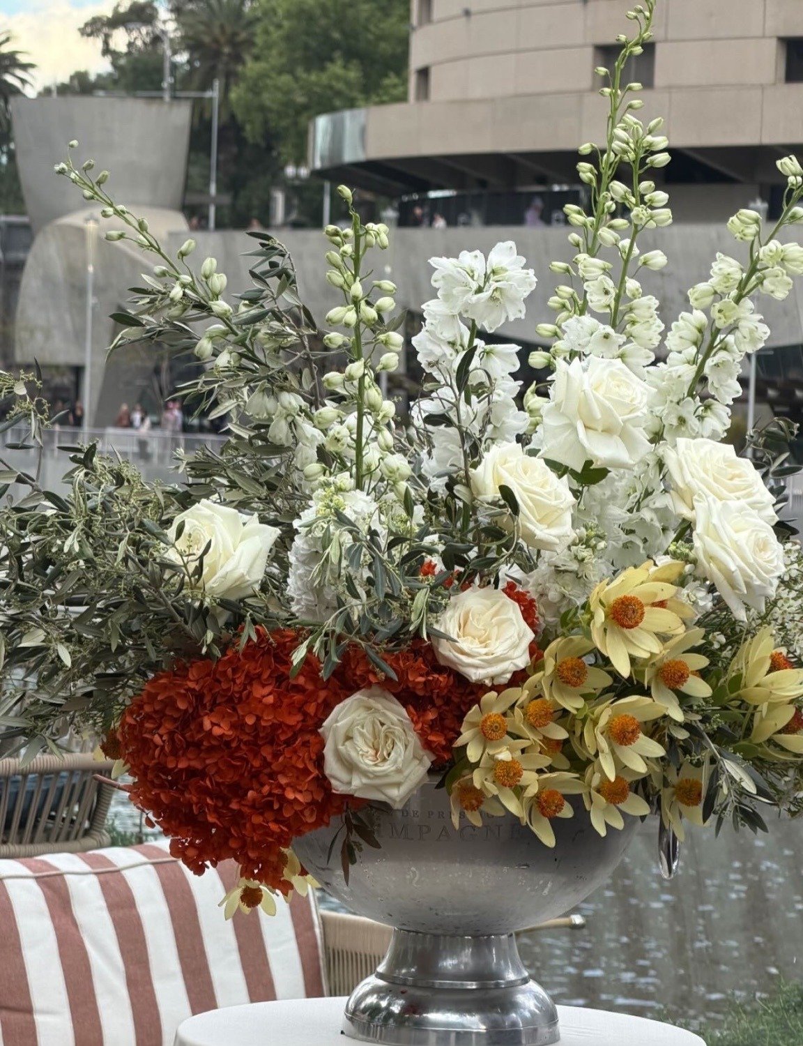 The orange, white and olive arrangement of roses, delphinium and hydrangea at Afloat Bar, with the Yarra River and the Melbourne skyline behind
