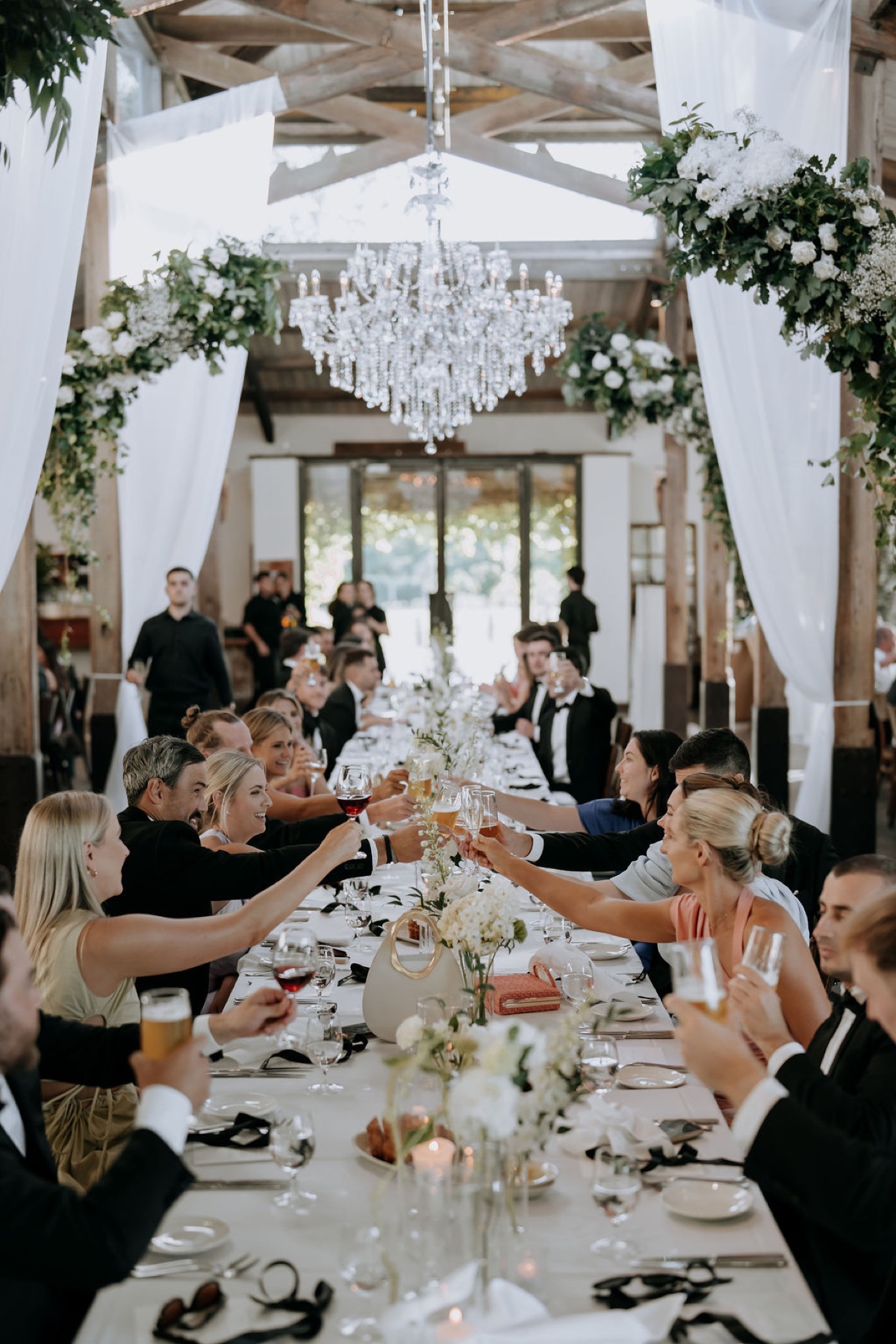 Wedding reception floral styling at Stones of the Yarra Valley, Victoria, Australia. White and green florals matched with a large Cinderella chandelier, drapings and hanging floral installations. Black ribbons on the tables for accent of contrast