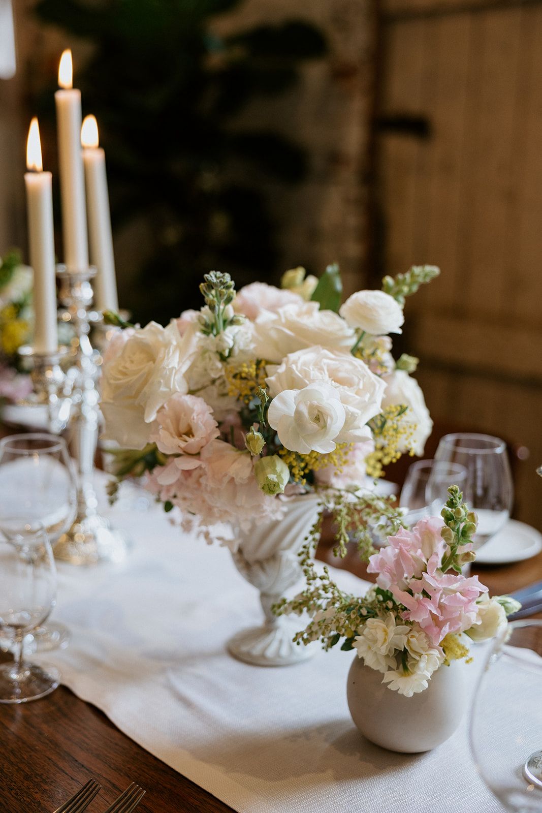 Fluted urn centrepiece of white roses, ranunculus and mimosa with taper candles at The Stables Restaurant
