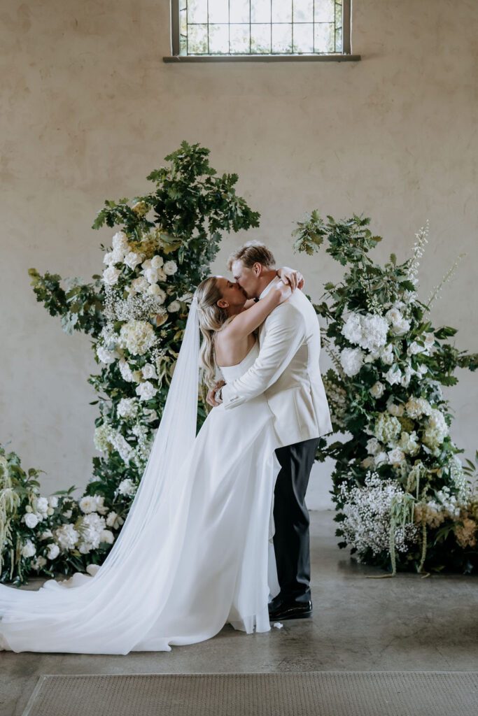 Bride and groom in front of a ceremony floral display of white and green. Lush oak foliage matched with roses, hydrangeas, delphinium and other premium white wedding florals.