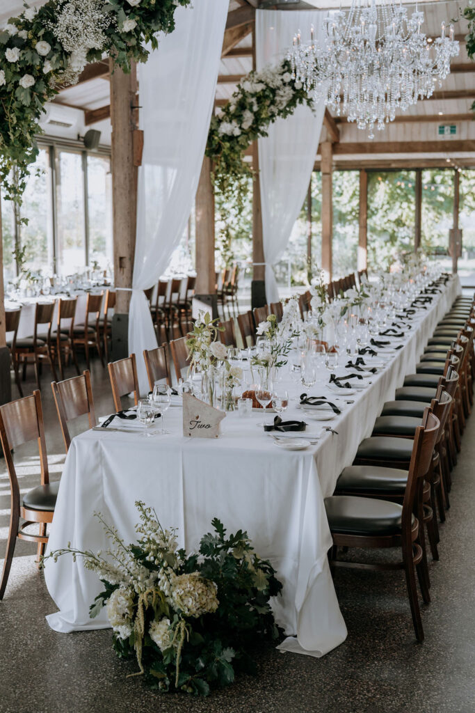 Wedding reception floral styling at Stones of the Yarra Valley, Victoria, Australia. White and green florals matched with a large Cinderella chandelier, drapings and hanging floral installations. Black ribbons on the tables for accent of contrast